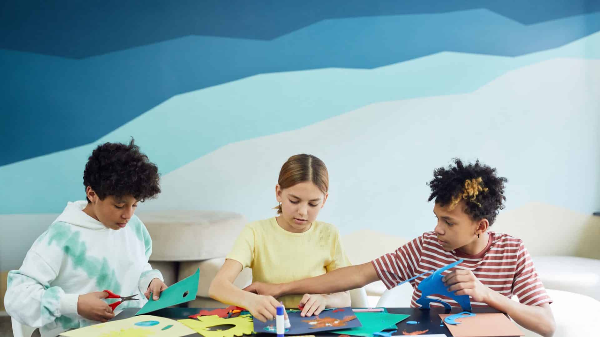 Trois enfants découpent du papier coloré autour d'une table d'atelier